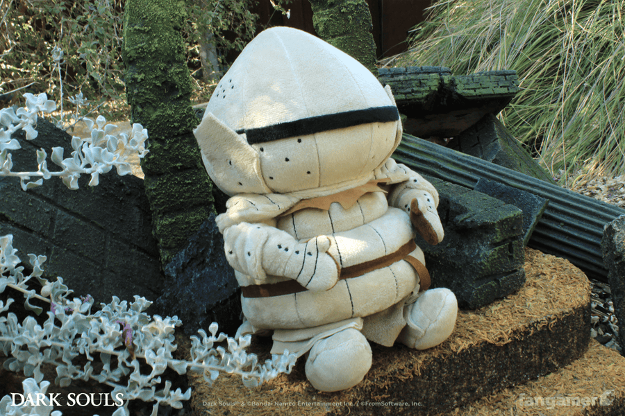 The Siegmeyer plush photographed outdoors at a 3/4 angle, seated next to a flowering bush and miniature ruins.  - Siegmeyer Plush - DARK SOULS