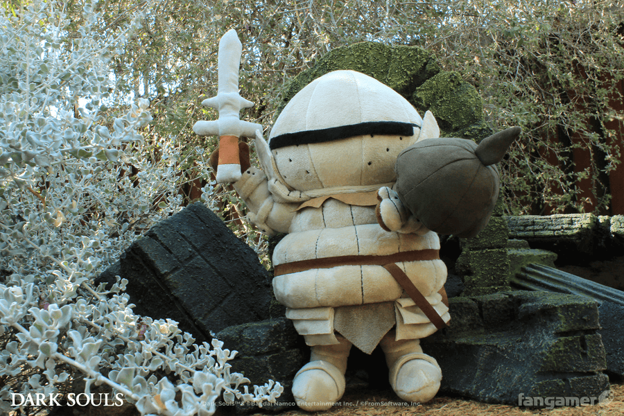 The Siegmeyer plush photographed outdoors, supported by the miniature ruins so that it appears to be standing and holding up its sword accessory and shield.  - Siegmeyer Plush - DARK SOULS